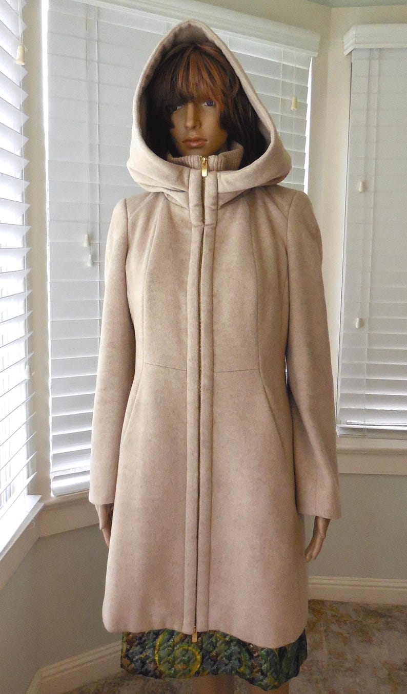 May include: A light beige hooded coat with a full-length zipper. The coat has a tailored fit and a hood that frames the face. The coat is displayed on a mannequin. The hemline of the coat is visible, with a patterned fabric.