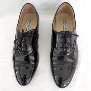 Puede incluir: Un par de zapatos Oxford negros de charol con un acabado brillante. Los zapatos tienen un dise&ntilde;o cl&aacute;sico con cordones y un tac&oacute;n bajo. El interior de los zapatos tiene una etiqueta que dice "RODOLFO VALENTINO".