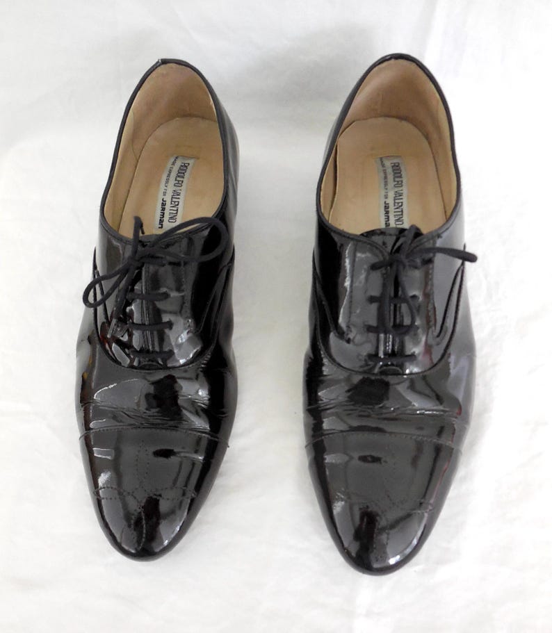 Puede incluir: Un par de zapatos Oxford negros de charol con un acabado brillante. Los zapatos tienen un dise&ntilde;o cl&aacute;sico con cordones y un tac&oacute;n bajo. El interior de los zapatos tiene una etiqueta que dice "RODOLFO VALENTINO".