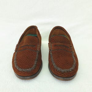 May include: A pair of brown loafers featuring a textured, pebbled surface and a classic penny slot strap across the vamp. The shoes have a moccasin-style stitched toe seam, a dark brown sole, and a visible green interior lining at the heel area.