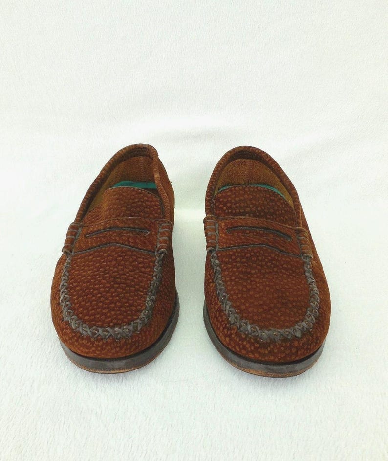May include: A pair of brown loafers featuring a textured, pebbled surface and a classic penny slot strap across the vamp. The shoes have a moccasin-style stitched toe seam, a dark brown sole, and a visible green interior lining at the heel area.