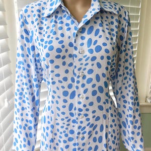 May include: A white shirt dress featuring a pattern of blue polka dots. The long-sleeved dress has a collared neckline and clear buttons down the front. The fabric appears to be lightweight.