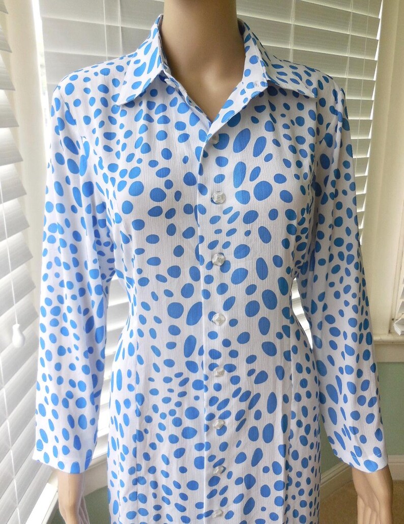 May include: A white shirt dress featuring a pattern of blue polka dots. The long-sleeved dress has a collared neckline and clear buttons down the front. The fabric appears to be lightweight.