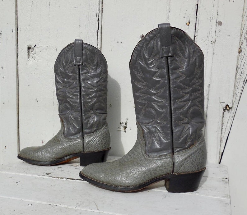 May include: A pair of gray cowboy boots. The boots have a textured gray foot and a smooth gray shaft with decorative stitching. The heels are black.