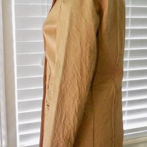 May include: A tan leather jacket with a long, fitted silhouette and a collar. The jacket has a textured surface and a subtle sheen.
