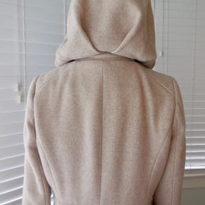 May include: A beige hooded coat, viewed from the back, showcasing a tailored design. The coat features a structured hood and a classic silhouette, suitable for outerwear. The fabric appears to have a subtle texture.