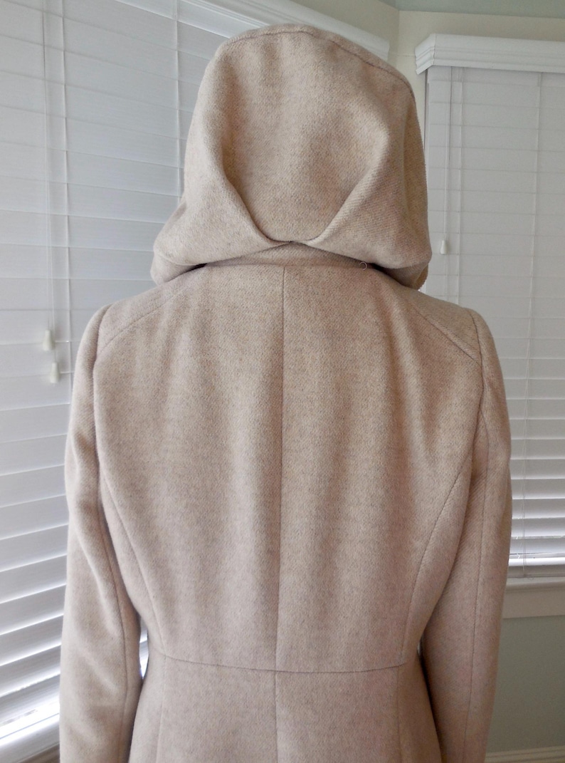 May include: A beige hooded coat, viewed from the back, showcasing a tailored design. The coat features a structured hood and a classic silhouette, suitable for outerwear. The fabric appears to have a subtle texture.