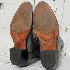 May include: Pair of worn gray cowboy boots, soles facing up. The soles are brown leather with visible wear and stitching. The heels are black, and the boots are marked with the number "21-Y". The boots are set against a white, weathered background.