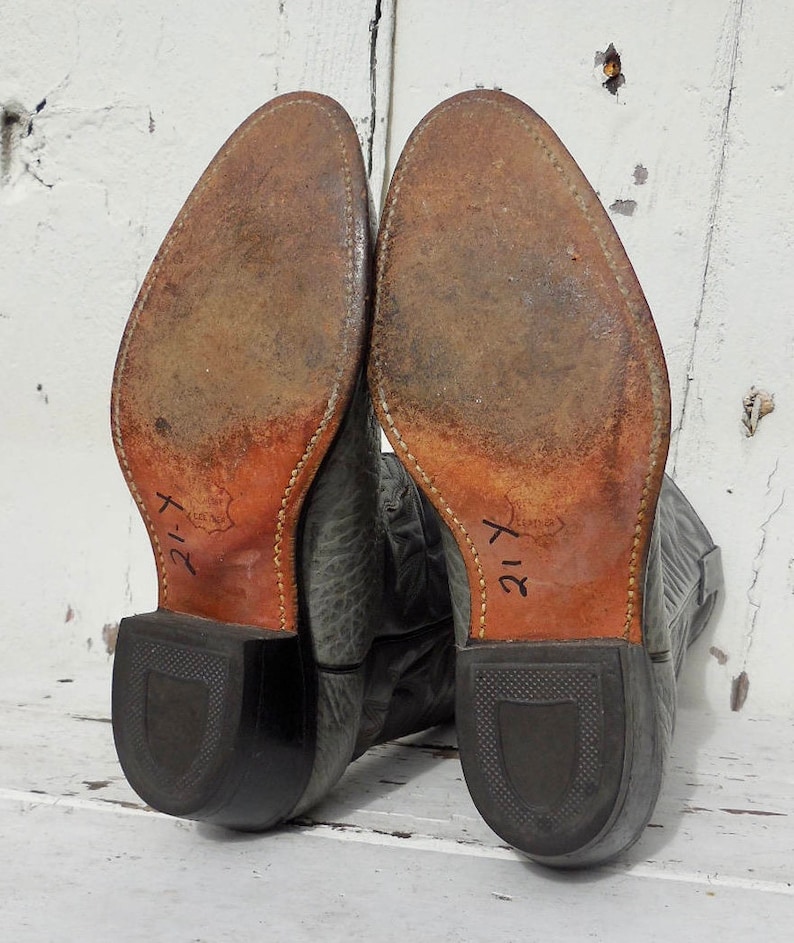 May include: Pair of worn gray cowboy boots, soles facing up. The soles are brown leather with visible wear and stitching. The heels are black, and the boots are marked with the number "21-Y". The boots are set against a white, weathered background.