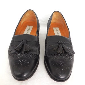 May include: Black leather tassel loafers with a woven pattern and perforated detailing. The loafers have a brown leather insole with the text "Murray's" printed on it.