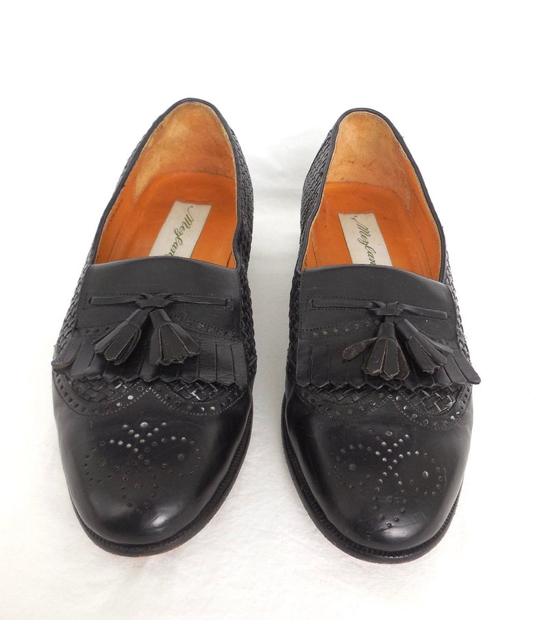 May include: Black leather tassel loafers with a woven pattern and perforated detailing. The loafers have a brown leather insole with the text "Murray's" printed on it.