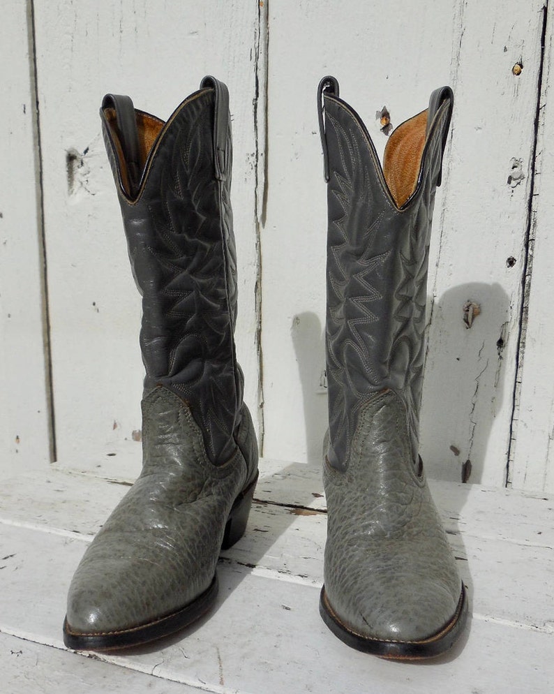 May include: A pair of gray cowboy boots with a textured finish on the lower part and smooth leather on the upper shaft. The boots have a pointed toe and a stacked heel, showcasing classic Western wear.