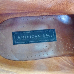 Pode incluir: Close-up do interior de um sapato de couro marrom com uma etiqueta preta que diz "AMERICAN RAG" em letras brancas.