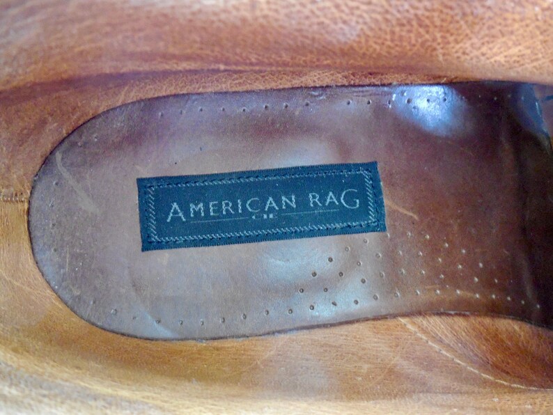 Pode incluir: Close-up do interior de um sapato de couro marrom com uma etiqueta preta que diz "AMERICAN RAG" em letras brancas.