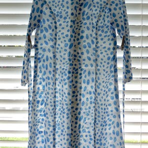 May include: A long, button-down dress with a white background and a pattern of blue oval shapes. The dress has long sleeves and a collar. The dress is hanging in front of a window with blinds.
