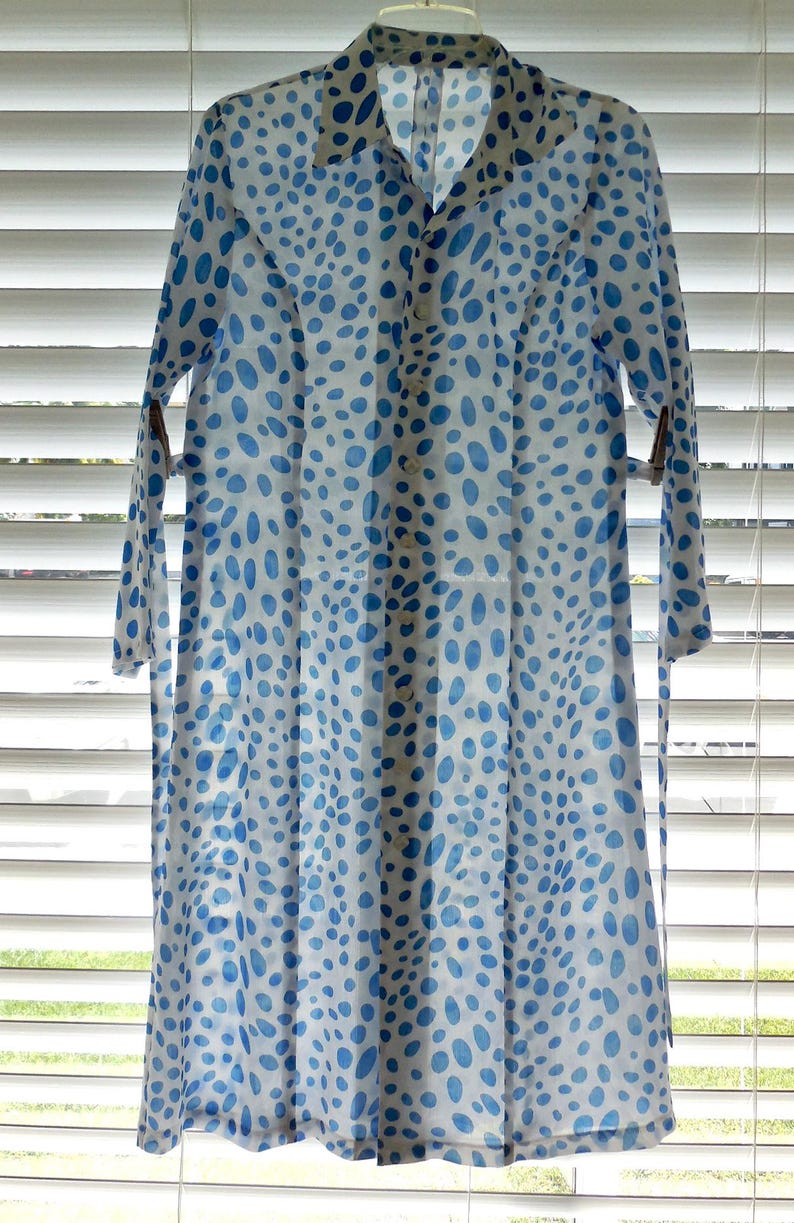 May include: A long, button-down dress with a white background and a pattern of blue oval shapes. The dress has long sleeves and a collar. The dress is hanging in front of a window with blinds.