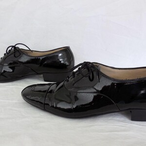 Puede incluir: Un par de zapatos de vestir negros de charol con acabado brillante. Los zapatos tienen un dise&ntilde;o Oxford cl&aacute;sico con puntera y cierre de cordones. Los tacones son bajos, sobre un fondo blanco.