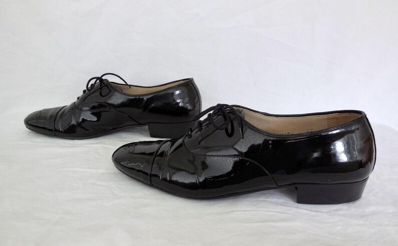 Puede incluir: Un par de zapatos de vestir negros de charol con acabado brillante. Los zapatos tienen un dise&ntilde;o Oxford cl&aacute;sico con puntera y cierre de cordones. Los tacones son bajos, sobre un fondo blanco.