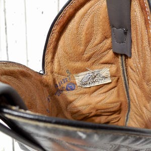 May include: Close-up of a black leather cowboy boot interior. The boot's interior is tan with intricate stitching and a Palomino USA label. A pull strap is visible, and the boot's size is marked inside.