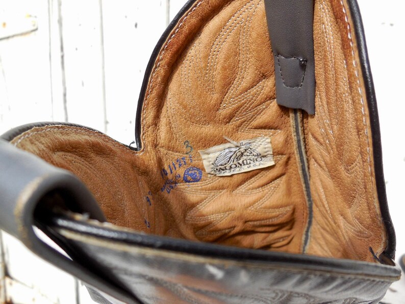 May include: Close-up of a black leather cowboy boot interior. The boot's interior is tan with intricate stitching and a Palomino USA label. A pull strap is visible, and the boot's size is marked inside.