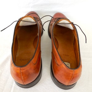 May include: A pair of brown leather dress shoes with dark brown laces. The shoes have a classic design with a rounded toe and a dark sole. The interior of the shoes is visible, showing a light-colored lining and the brand name.