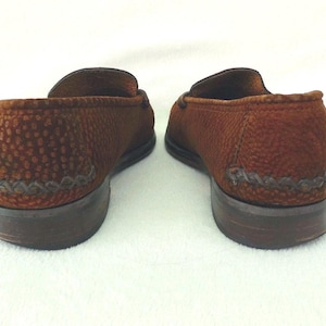 May include: A pair of brown loafers featuring a textured, pebbled surface and a decorative braided leather detail along the heel. The shoes have a low, stacked brown heel and a rounded toe silhouette, presented from a rear-facing perspective.