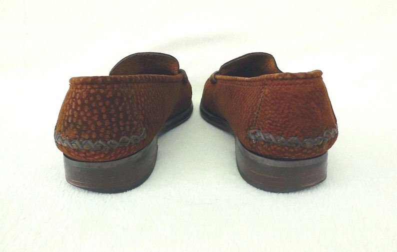 May include: A pair of brown loafers featuring a textured, pebbled surface and a decorative braided leather detail along the heel. The shoes have a low, stacked brown heel and a rounded toe silhouette, presented from a rear-facing perspective.