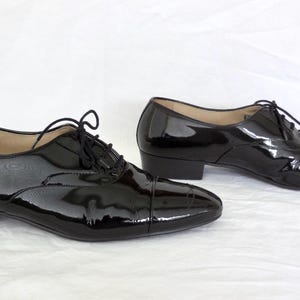 Puede incluir: Un par de zapatos Oxford de charol negro con tac&oacute;n bajo. Los zapatos tienen un dise&ntilde;o cl&aacute;sico con puntera y cierre de cordones. El acabado brillante refleja la luz. Fondo blanco.