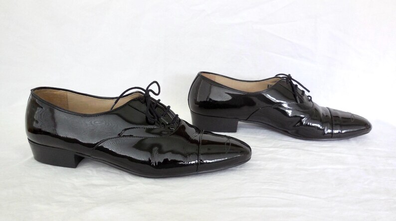 Puede incluir: Un par de zapatos Oxford de charol negro con tac&oacute;n bajo. Los zapatos tienen un dise&ntilde;o cl&aacute;sico con puntera y cierre de cordones. El acabado brillante refleja la luz. Fondo blanco.
