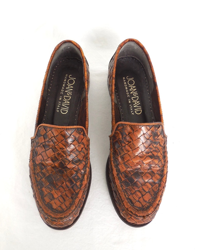 May include: A pair of brown leather loafers with a woven pattern. The loafers are handmade in Italy and have the brand name "Joan & David" printed on the inside.