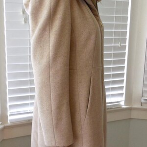 May include: A light beige hooded coat with a tailored fit. The coat features a hood, long sleeves, and a knee-length hem. The coat is made of a soft, textured fabric and has a classic, timeless design. A versatile outerwear piece.