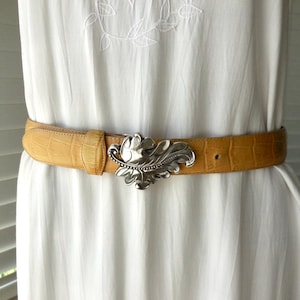 May include: A tan leather belt with a silver leaf-shaped buckle. The belt has a textured, crocodile-like pattern. The buckle is a detailed, ornate design. The belt is worn over a white dress.