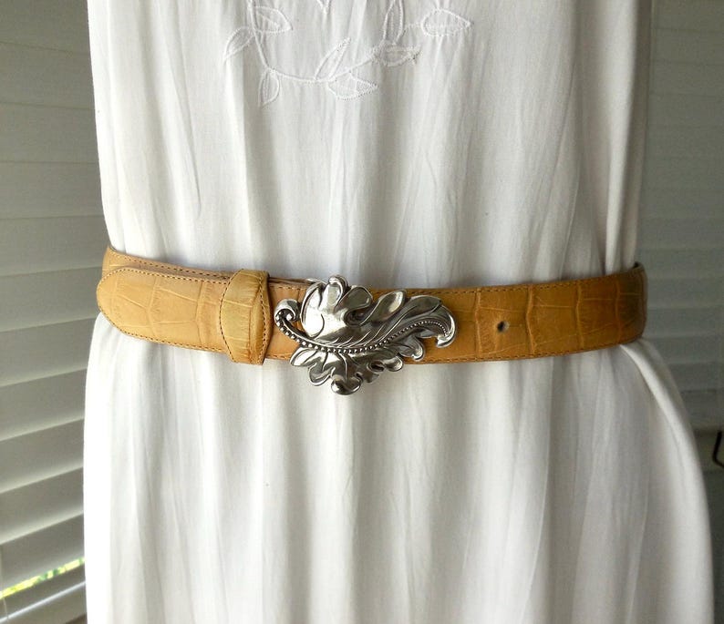 May include: A tan leather belt with a silver leaf-shaped buckle. The belt has a textured, crocodile-like pattern. The buckle is a detailed, ornate design. The belt is worn over a white dress.