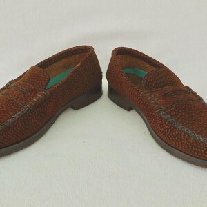 May include: A pair of brown loafers with a textured, pebbled surface and a classic penny slot strap across the vamp. The shoes feature a low heel, a rounded toe, and a visible teal-colored insole lining inside the opening of each shoe.