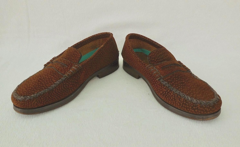 May include: A pair of brown loafers with a textured, pebbled surface and a classic penny slot strap across the vamp. The shoes feature a low heel, a rounded toe, and a visible teal-colored insole lining inside the opening of each shoe.