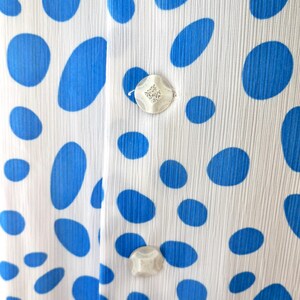 May include: Close-up of white fabric patterned with large blue ovals. The fabric has a vertical ribbed texture and is fastened with two decorative, iridescent buttons.