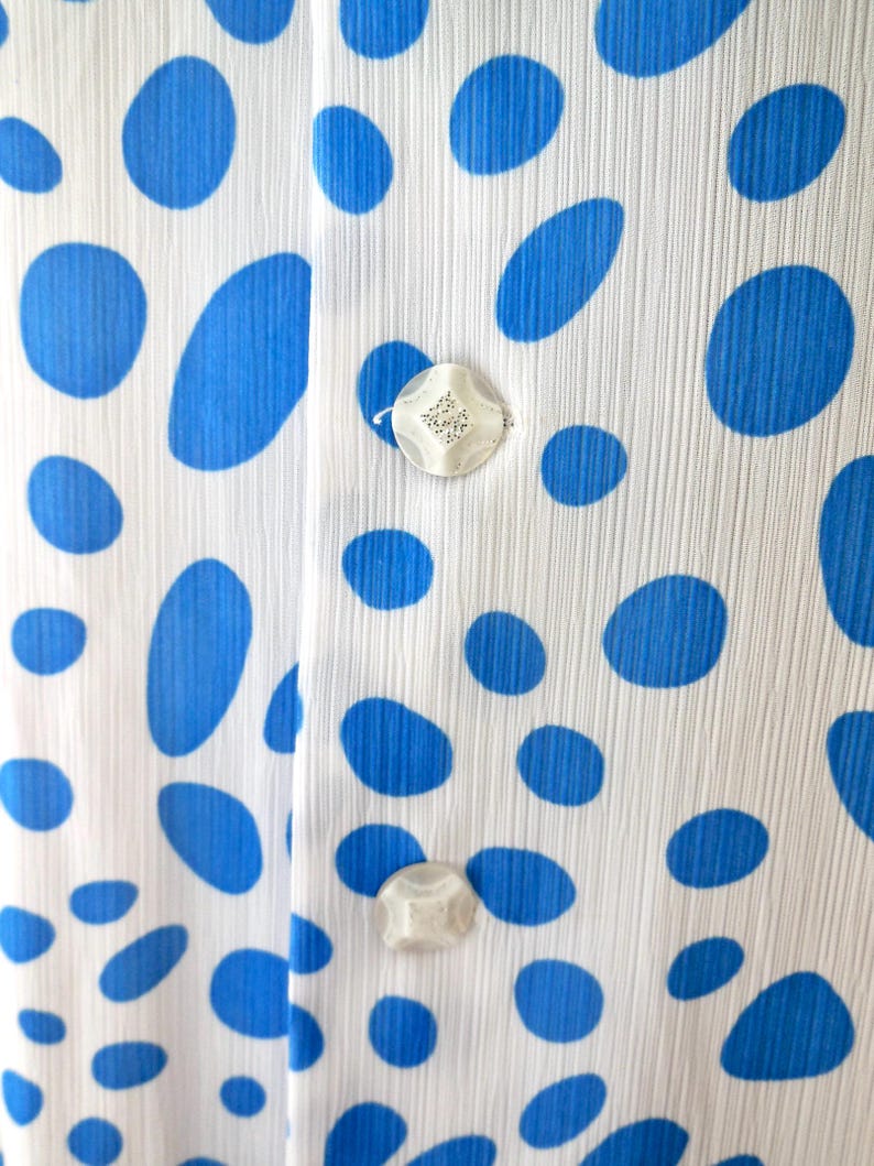 May include: Close-up of white fabric patterned with large blue ovals. The fabric has a vertical ribbed texture and is fastened with two decorative, iridescent buttons.