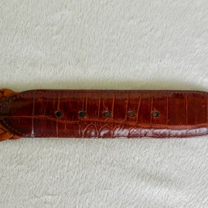 May include: A brown leather belt with a woven design and a smooth leather buckle. The buckle has five holes for adjusting the fit.