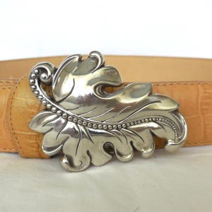 May include: Tan leather belt featuring a silver leaf-shaped buckle. The belt has a textured, crocodile-style pattern. The buckle is detailed with a beaded edge and leaf veins. A fashionable accessory.