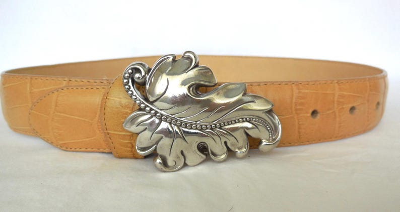 May include: Tan leather belt featuring a silver leaf-shaped buckle. The belt has a textured, crocodile-style pattern. The buckle is detailed with a beaded edge and leaf veins. A fashionable accessory.