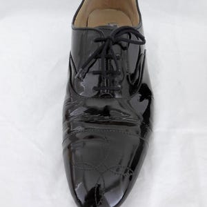Puede incluir: Zapato Oxford de charol negro con acabado brillante. El zapato presenta cierre de cordones y costuras decorativas en la puntera. Adecuado para ocasiones formales o elegantes.