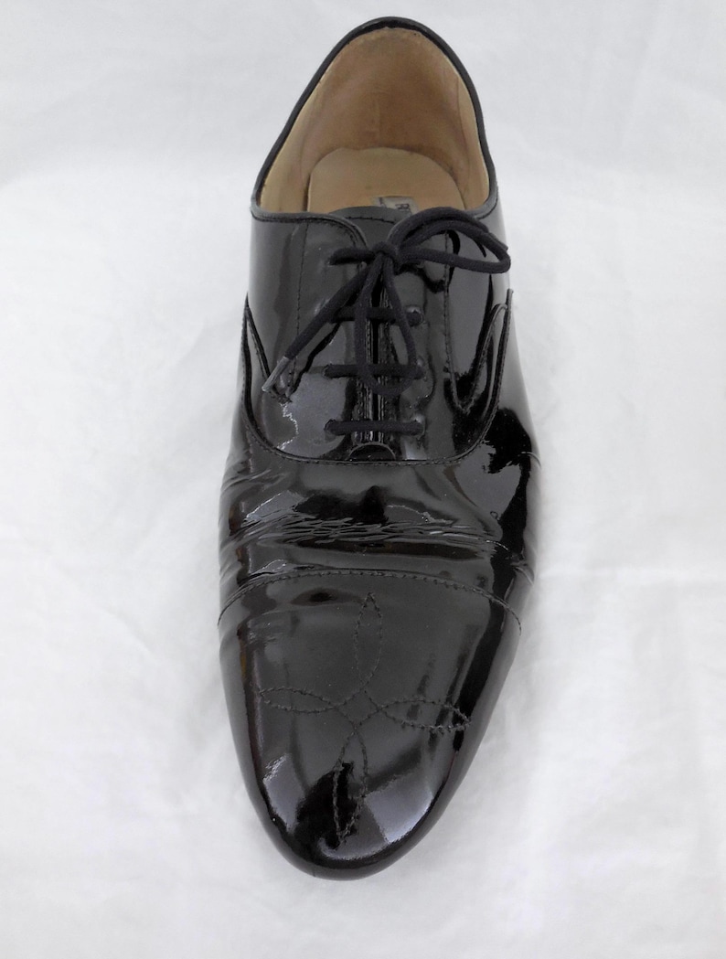 Puede incluir: Zapato Oxford de charol negro con acabado brillante. El zapato presenta cierre de cordones y costuras decorativas en la puntera. Adecuado para ocasiones formales o elegantes.