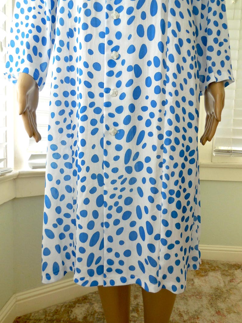 May include: A white dress featuring a blue polka dot design. The dress has long sleeves and a button-up front. The knee-length dress is made of a lightweight material, suitable for warmer weather.