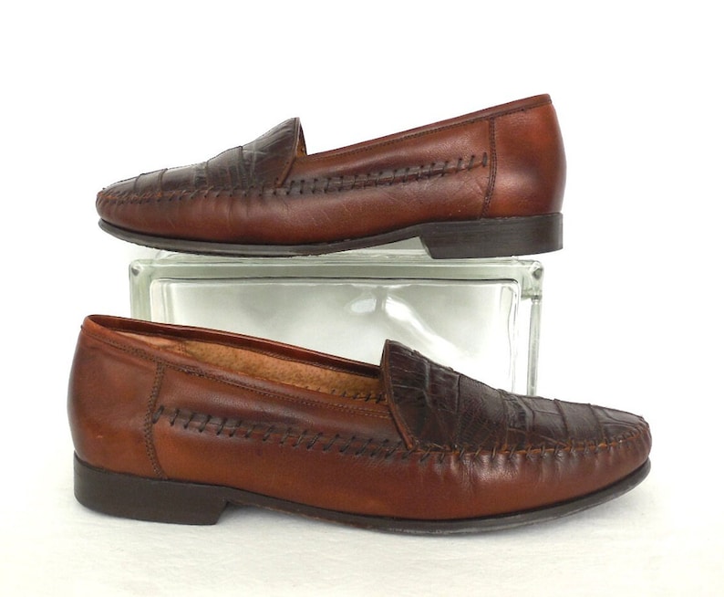 K&ouml;nnte beinhalten: Ein Paar braune Lederschuhe mit Kroko-Pr&auml;gung an der Spitze. Die Schuhe haben ein gesticktes Detail am Rand und eine braune Ledersohle.