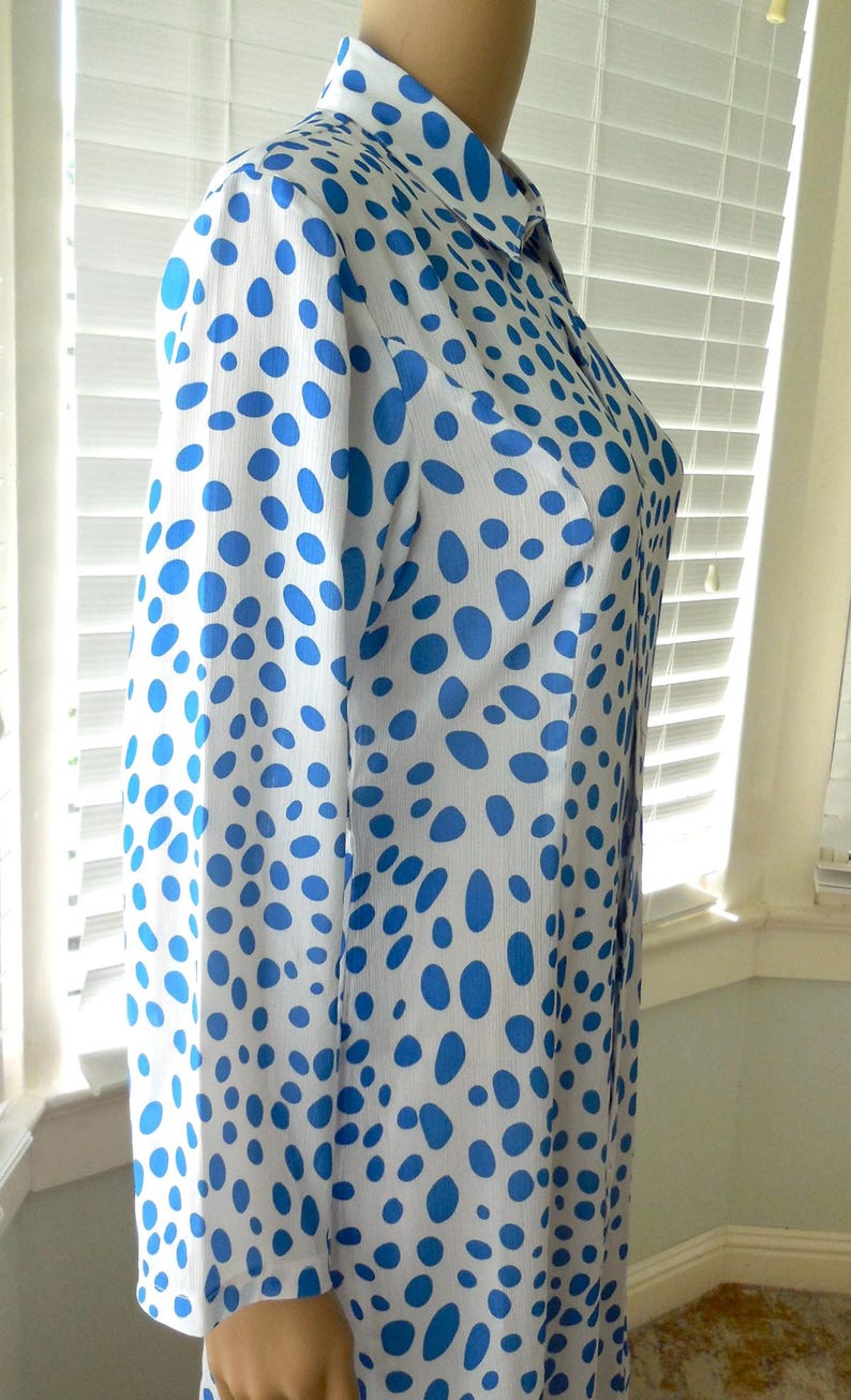 May include: A white dress featuring a blue polka dot design. The dress has long sleeves and a collared neckline. The fabric appears lightweight, and the dress is buttoned down the front. The dress is displayed on a mannequin.