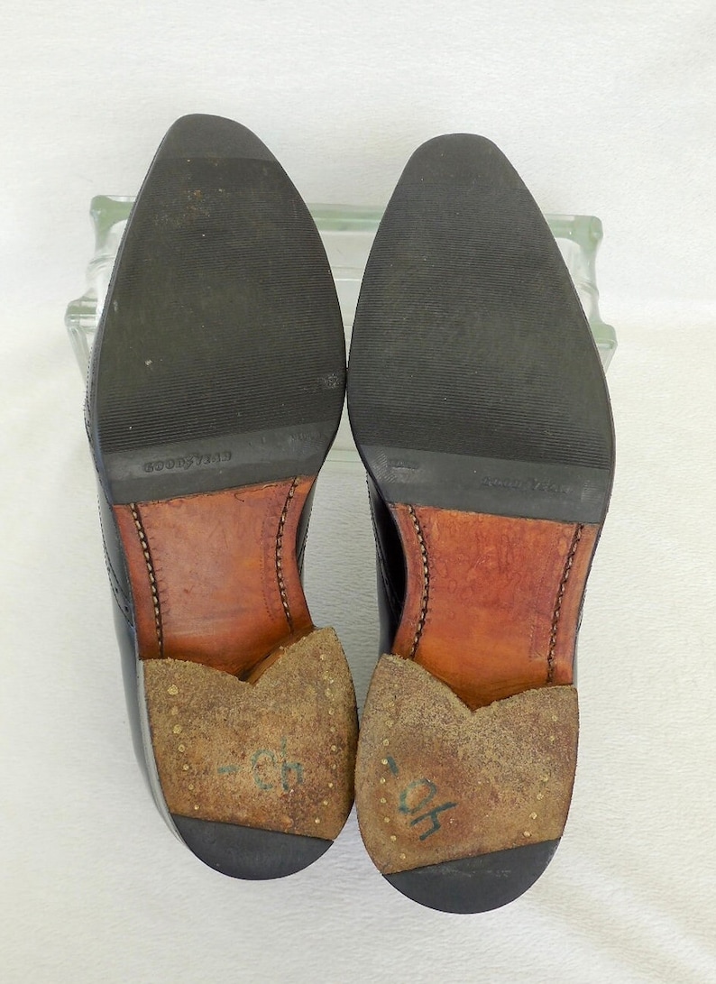 Puede incluir: Primer plano de las suelas de dos zapatos de cuero negro. Los zapatos tienen plantillas de cuero marr&oacute;n y est&aacute;n marcados con el texto "GOOD YEAR" y "GOODYEAR".