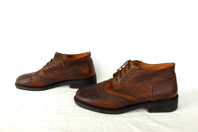 Pode incluir: Um par de botas de tornozelo de couro marrom com fecho de cadar&ccedil;o. As botas t&ecirc;m um padr&atilde;o decorativo de pequenos furos e costuras.