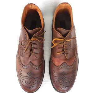 Pode incluir: Um par de sapatos de couro marrom com brogues e cadar&ccedil;os. Os sapatos t&ecirc;m um padr&atilde;o decorativo de pequenos furos e costuras.