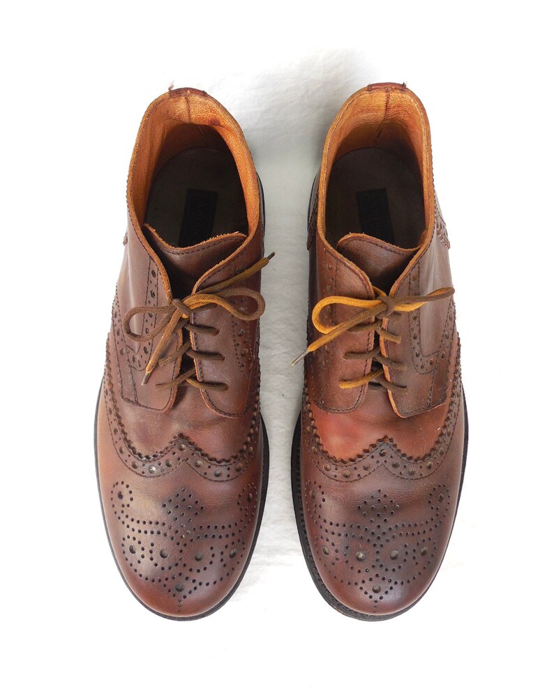 Pode incluir: Um par de sapatos de couro marrom com brogues e cadar&ccedil;os. Os sapatos t&ecirc;m um padr&atilde;o decorativo de pequenos furos e costuras.