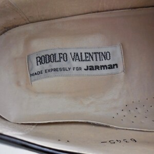 Puede incluir: Vista interior de un zapato negro con una plantilla color crema. Una etiqueta rectangular dice "RODOLFO VALENTINO MADE EXPRESSLY FOR JARMAN" en texto negro. La plantilla del zapato tiene peque&ntilde;os agujeros de ventilaci&oacute;n.